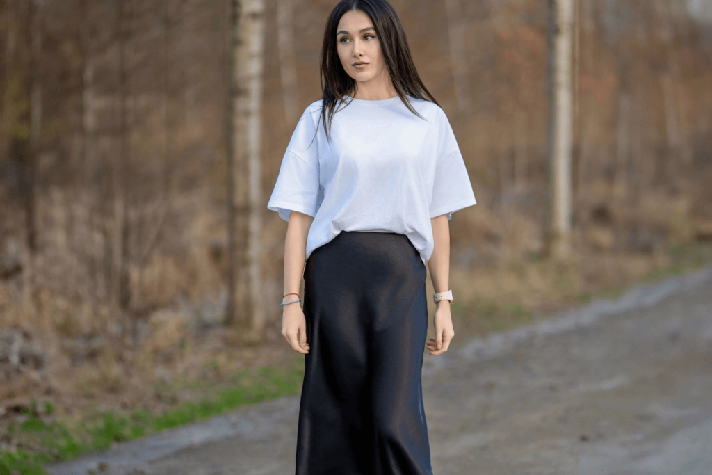 A woman in a white oversized t-shirt and a black mid-length pencil skirt, standing outdoors with a serene expression, surrounded by nature and walking path.