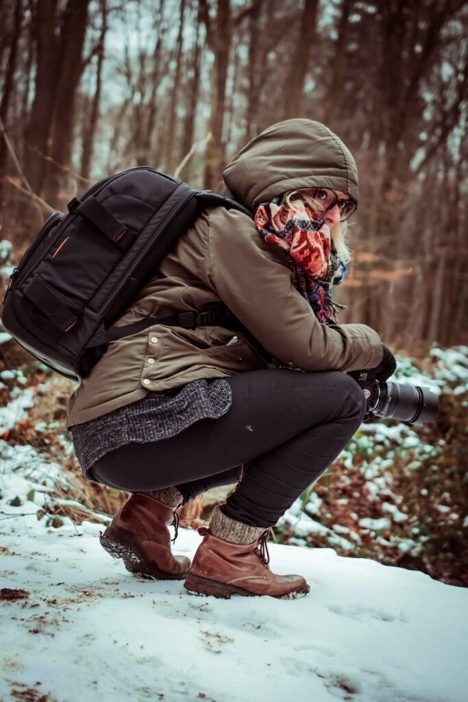 A photographer in an olive-green winter jacket, patterned scarf, and brown leather boots crouching on snow, holding a camera with a telephoto lens in a forest setting.