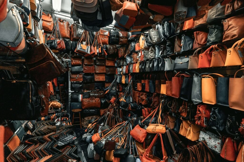 A shop or storage room filled with a large variety of leather bags and purses in different colors, styles, and sizes. The shelves are stacked from floor to ceiling, and additional bags are hanging and piled on the floor, creating a cluttered yet vibrant scene.