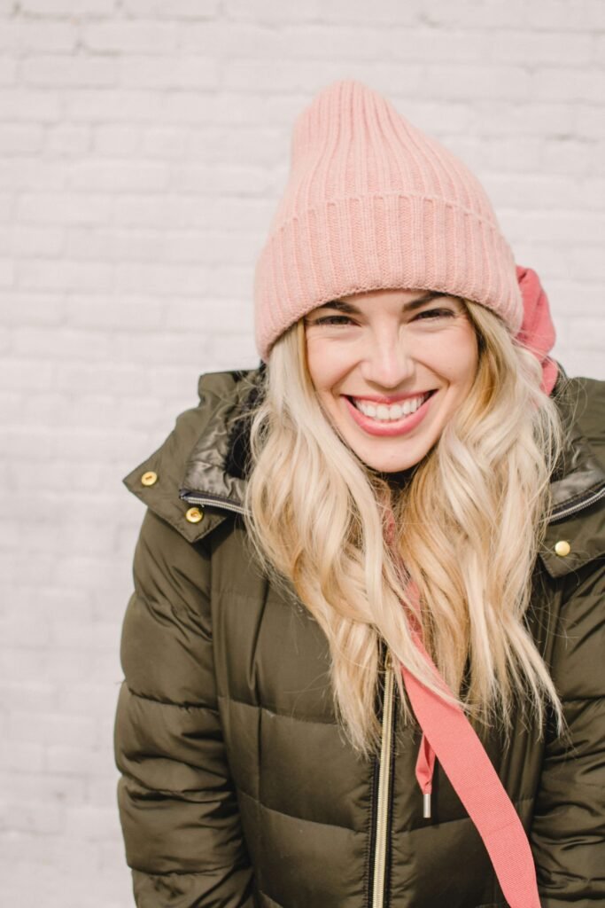 A smiling woman wearing a pink knitted beanie and an olive-green winter jacket, perfect for extreme cold weather, against a light brick background.