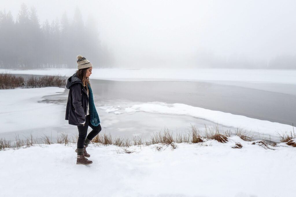 A person walking along a snowy lakeshore, wearing a winter coat, knitted hat, and boots, with a misty background and frozen lake.