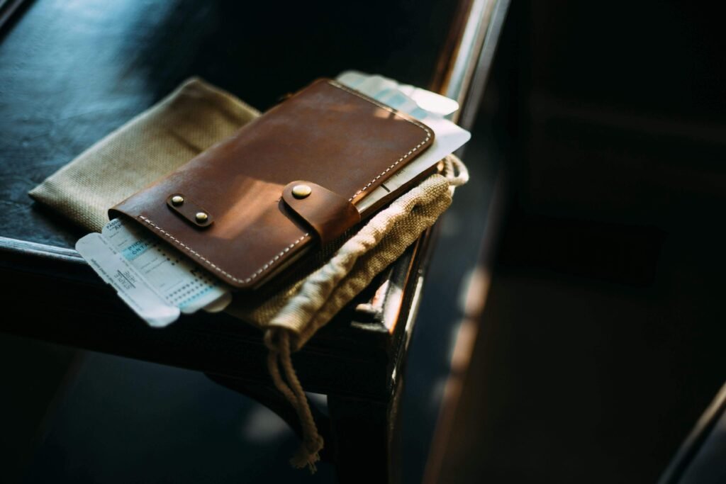 A close-up of a stylish brown leather wallet resting on a wooden table, with travel tickets partially visible inside. The wallet is placed on a beige fabric pouch, creating a warm and vintage aesthetic.