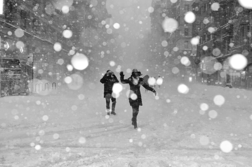 Two people are walking down a snow-covered street, both wearing lightweight winter jackets with hoods. Snowflakes are gently falling around them, and their jackets provide light protection against the cold as they stroll through the wintery scene. The surrounding area is blanketed in snow, creating a peaceful and chilly atmosphere.