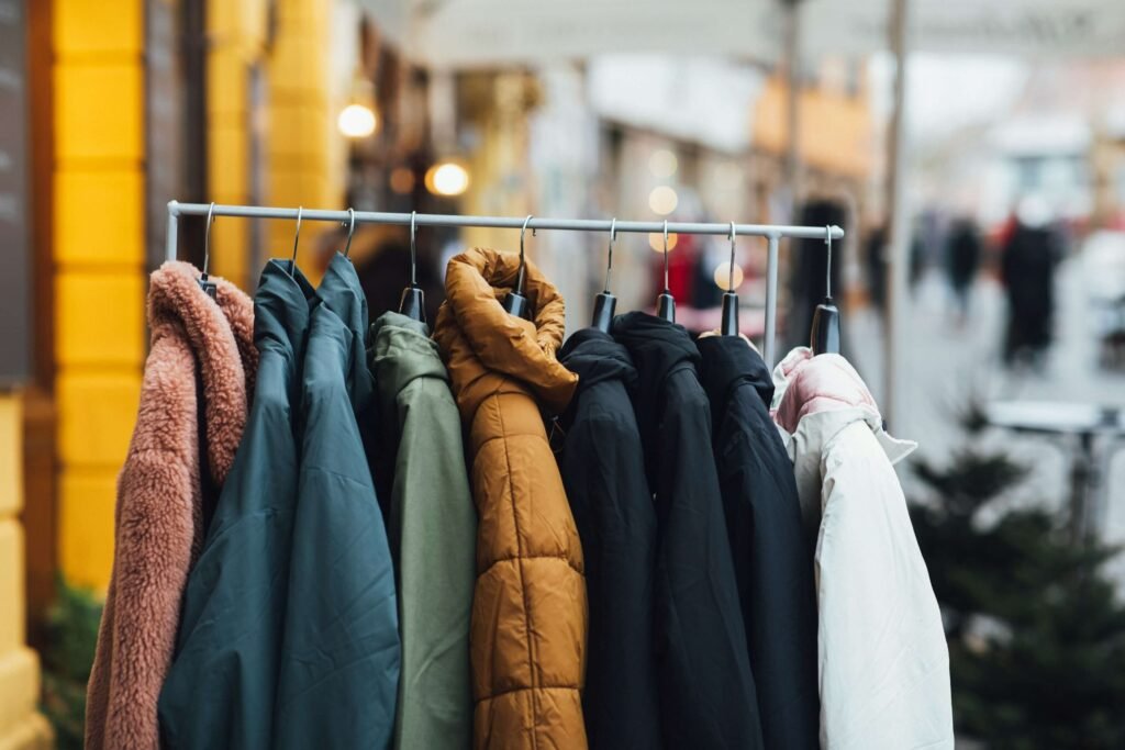 A rack of lightweight winter jackets in various colors, including green, brown, black, pink, and beige, displayed outdoors on a blurred street background.