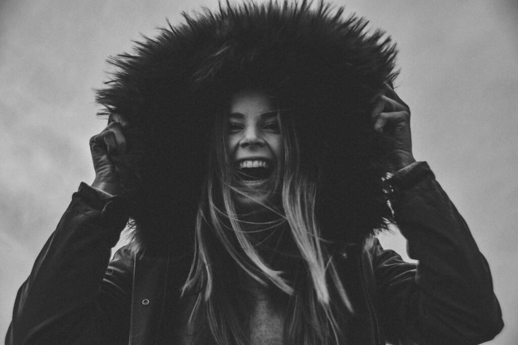 A close-up of a young woman smiling, wearing a women's coat with the hood up.